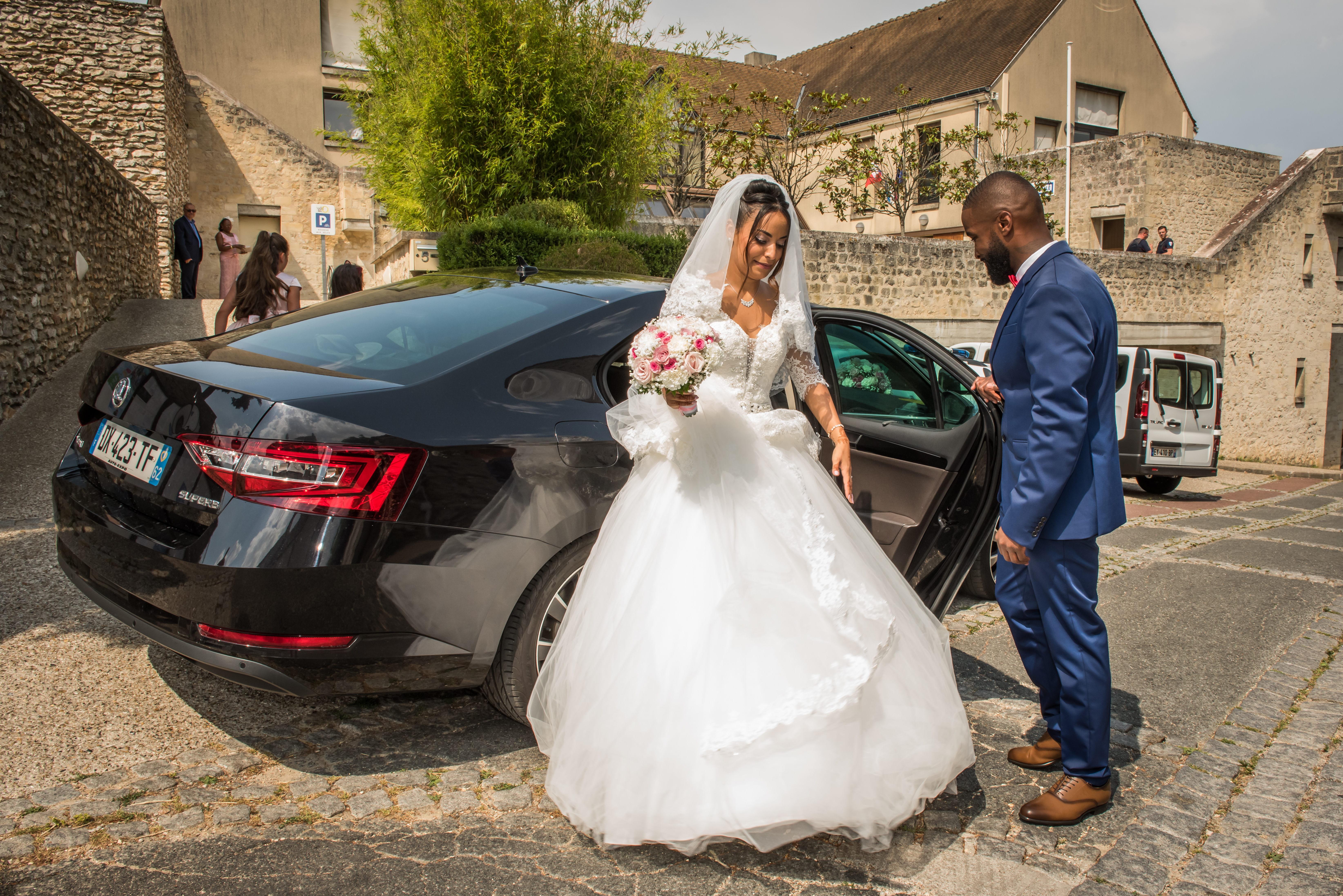 Mariage de Zara et Christy cérémonie arrivée à la mairie, le marié, en costume bleu, aide la marié, en robe blanche, à sortir de la voiture