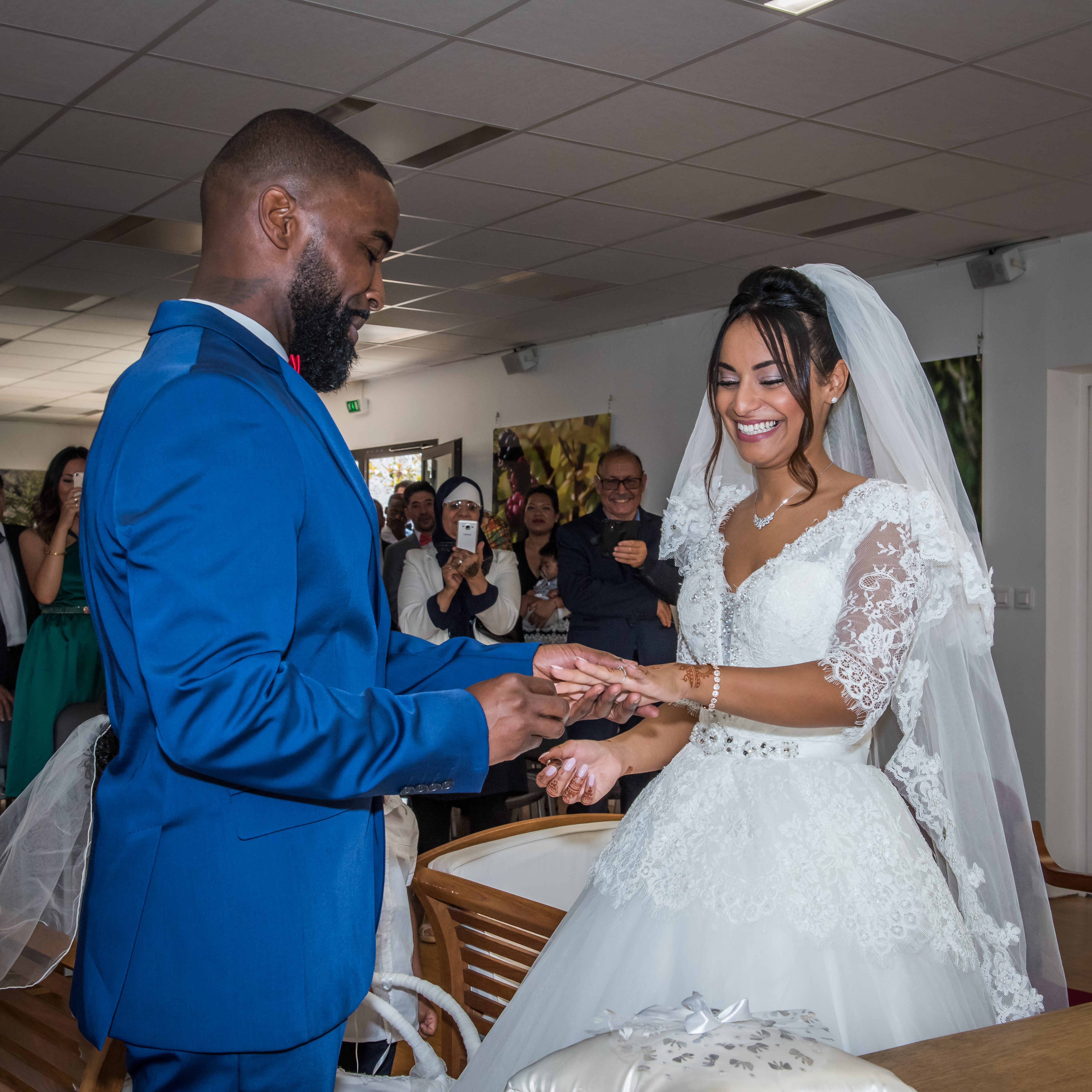 Mariage de Zara et Christy cérémonie échange d'alliance, le marié, en costume bleu, passe l'alliance au doigt de la mariée, en robe et voile blanc, devant les invités qui les prennent en photo