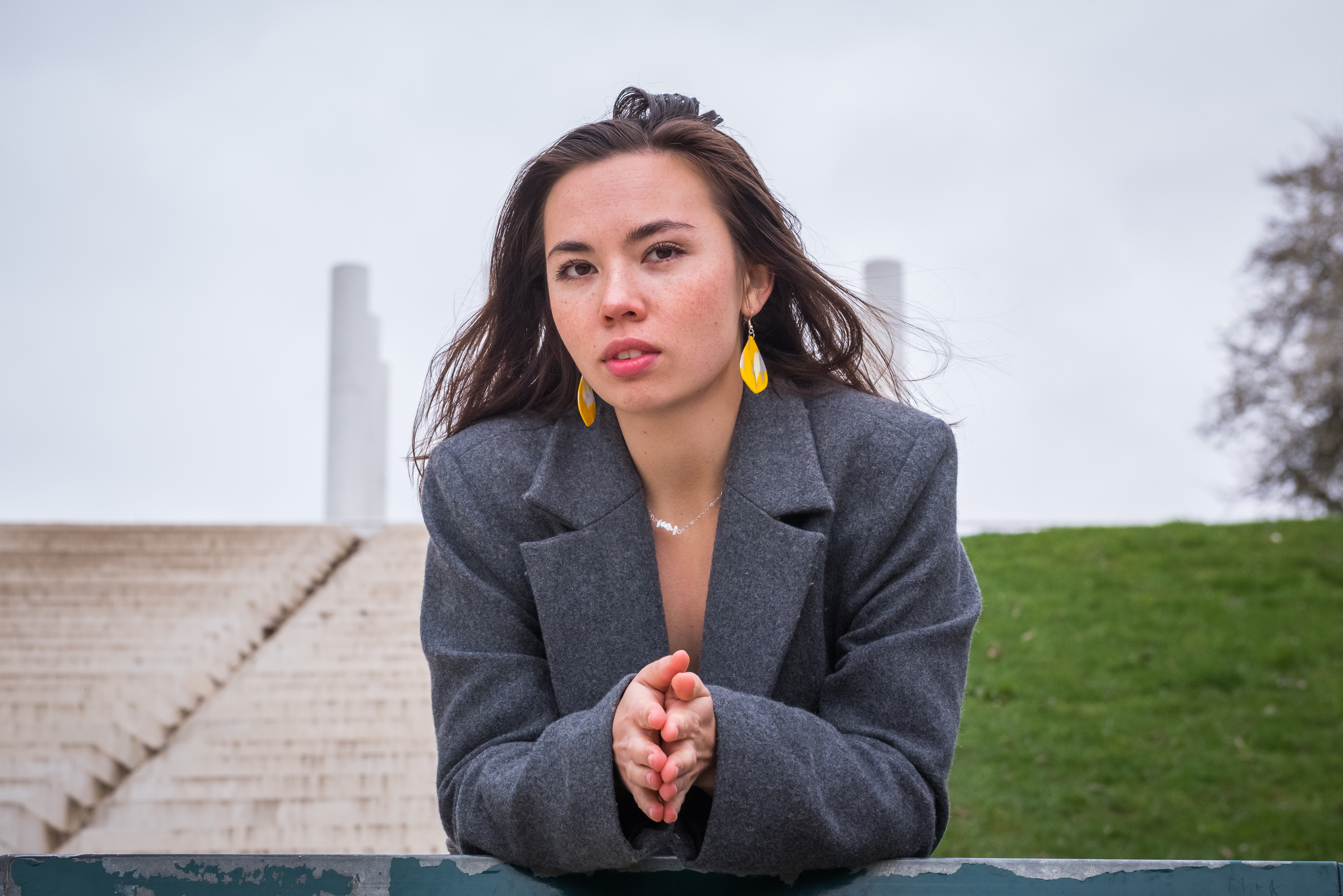 Shooting photo portrait solo en milieu urbain. Photo en plan taille, modèle adossée à une rambarde. La modèle porte un manteau long gris, de grandes boucles d'oreilles jaunes et un collier en pierres blanches.