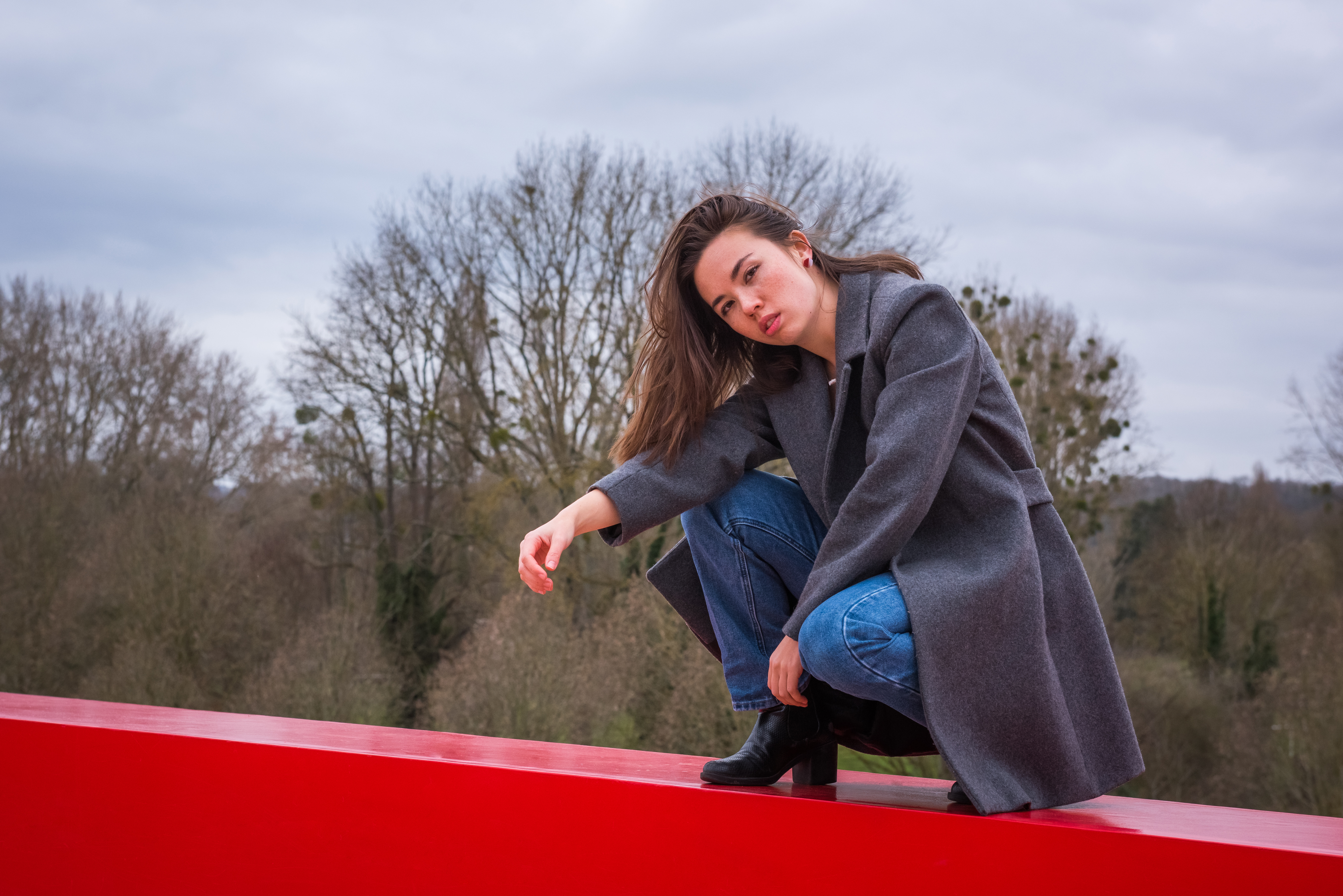 Shooting photo portrait solo en milieu urbain. Photo en pieds, modèle accroupi sur un muret, regarde l'objectif. La modèle porte un jean bleu, un t-shirt noir, un manteau long gris et de petites boucles d'oreilles rouge et blanche.